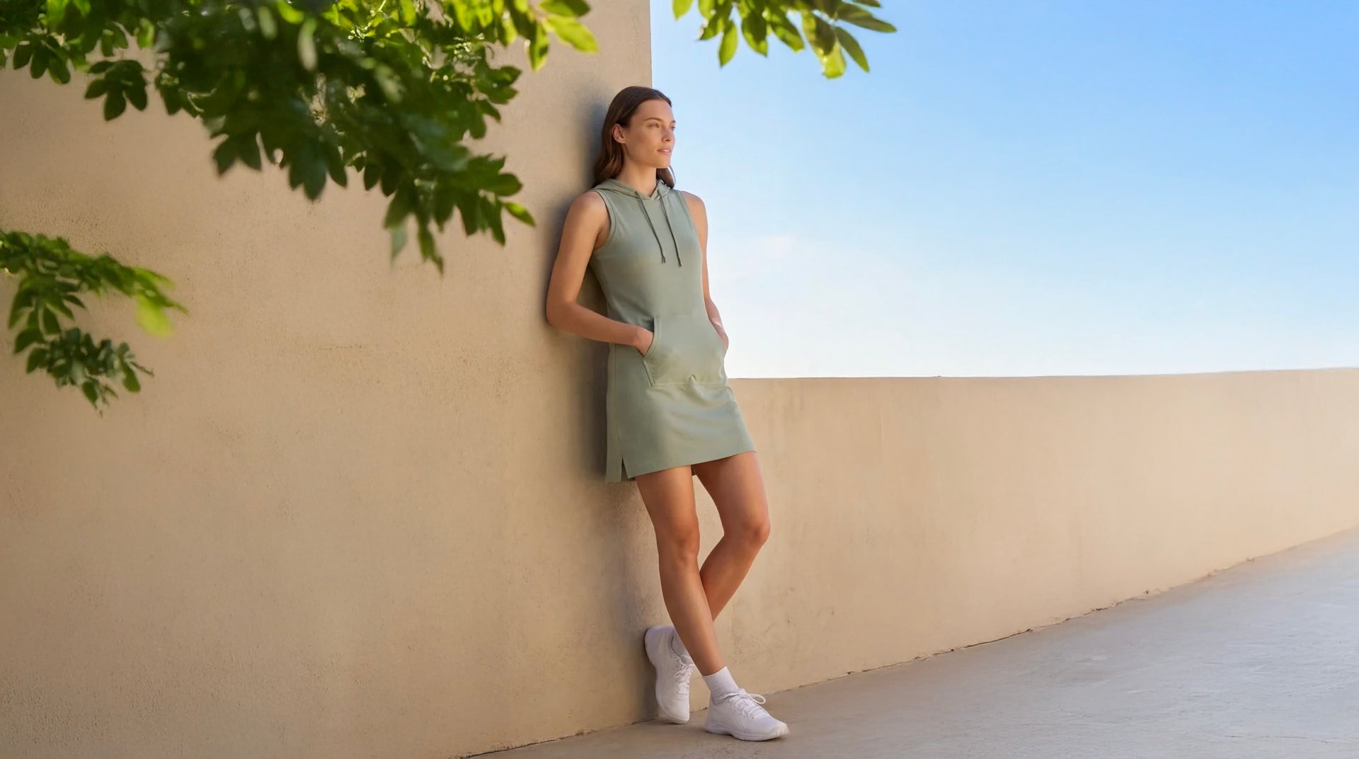 Woman in a green dress leaning against a beige wall with greenery and blue sky in the background